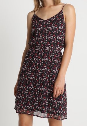 Vrouw draagt een mouwloze zwarte jurk met kleine rode, roze en witte bloemenpatronen en dunne schouderbandjes, staand tegen een effen achtergrond.