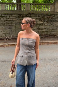 Top bez ramienok s kockovaným vzorom, gombíkovou prednou časťou, dvoma vreckami a priliehavým strihom; skombinovaný s voľnými džínsami. Drží tkanú tašku.