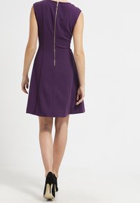 Robe sans manches violette avec un corsage ajusté et une jupe évasée, dotée d'une fermeture éclair dorée au dos, associée à des talons noirs.