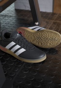 Black athletic shoes with three white stripes, textured upper, rubber sole with a gum finish, and padded tongue. Low-profile design.