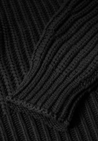 Maglione nero lavorato a maglia con texture a costine, con un primo piano sull'area dei polsi che evidenzia il filato spesso e il motivo distintivo.