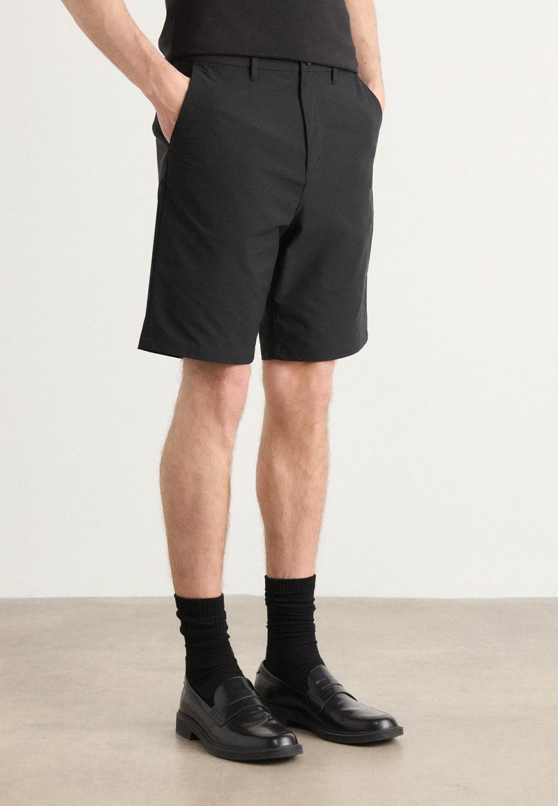Svarte shorts laget av glatt stoff, med sidelommer og en skreddersydd passform. Kombinert med svarte sokker og loafers, som skaper et enhetlig utseende.
