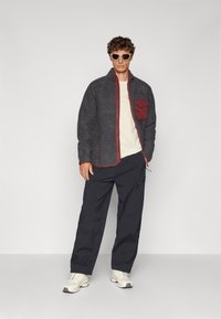 Fleeceová bunda v tmavě šedé barvě s vínovou kapsou, kombinovaná s námořnickými kalhotami a bílými teniskami. Model má na sobě bílé sluneční brýle.