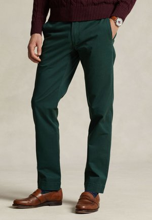 Homme portant un pantalon vert foncé, des mocassins en cuir marron, un pull bordeaux et une montre-bracelet avec un bracelet en cuir marron.