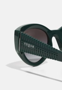 Zelene plastične sončne očala s sodobnim okvirjem, okroglimi lečami in teksturiranimi zelenimi ročaji, ki prikazujejo srebrni logotip "vogue".