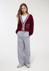 Giovane donna in piedi, indossa un cardigan bordeaux abbottonato con strisce bianche e toppa con la lettera "K", pantaloni ampi grigi e sneakers argento.