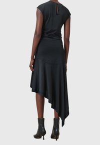 En kvinna bär en svart asymmetrisk midiklänning med en matchande svart topp och svarta klackboots vid anklarna, stående vänd bort.