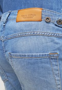Jeans en denim bleu clair avec une texture lisse, présentant un patch de marque en cuir marron et des coutures visibles au niveau de la poche arrière.
