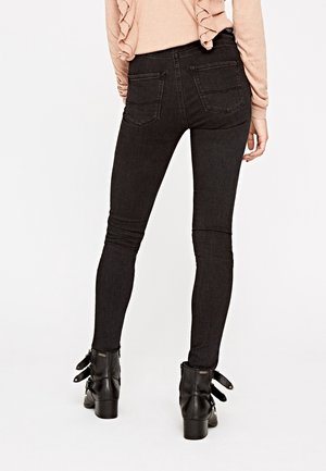 Persoon die zwarte skinny jeans met achterzakken draagt, een lichtbeige top met lange mouwen en ruches en zwarte enkellaarsjes met gespen.