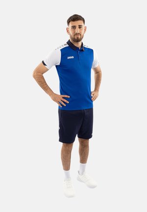 Mann steht mit den Händen in der Hüfte, trägt ein blau-weißes Polo, marineblaue Shorts, weiße Socken und weiße Turnschuhe vor einem einfarbigen Hintergrund.