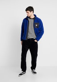 Jachetă din fleece albastră cu accente negre, cu fermoar frontal și buzunar pe piept. Pur și simplu peste o cămașă cu dungi alb-negru și pantaloni din corduroy negri.