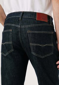 Tmavomodré džínsy z denimu s rovnými nohavicami, prešívanými zadnými vreckami a hnedou koženou nášivkou s logom na páse.