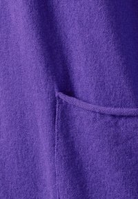 Lila gestrickter Stoff mit einer weichen Textur und einem kleinen Taschendetail. Die Nähte sind entlang der Kanten der Tasche sichtbar.