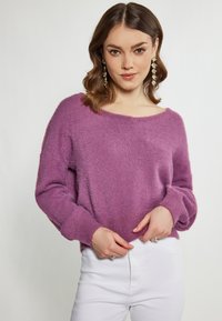 Lila, bolyhos kötött pulóver, széles nyakkivágással és hosszú ujjakkal, fehér, magas derekú nadrággal párosítva. Bordázott mandzsettákkal és szegéllyel rendelkezik.