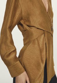 Top avvolgente marrone realizzato in tessuto morbido, con scollatura a V profonda, maniche lunghe e chiusura con fiocco laterale per una silhouette aderente.