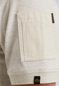 Chemise beige à manches courtes avec un tissu texturé, une poche plaquée avec des coutures détaillées et une petite étiquette logo noire et dorée.