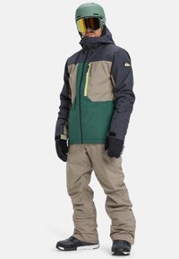 Mann in einer grünen, beigen und dunkelgrauen Snowboardjacke, beigen Schneehosen, schwarzen Handschuhen, einem grünen Helm und reflektierenden Goggles, stehend.