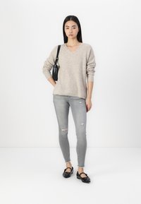 Beiger Strick-V-Ausschnitt-Pullover, hellgraue, verwitterte Skinny-Jeans und schwarze flache Schuhe mit silbernen Akzenten, vor einem schlichten Hintergrund stehend.