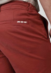 Pantalons en coton rouge avec une poche arrière portant une étiquette blanche avec trois silhouettes de vaches. Le tissu a une texture lisse et une coupe légèrement cintrée.