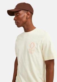 Lyscreme bomulds t-shirt med korte ærmer, prydet med et logo design i blød orange. Brun kasket med buet skygge; begge er afslappede stilarter.