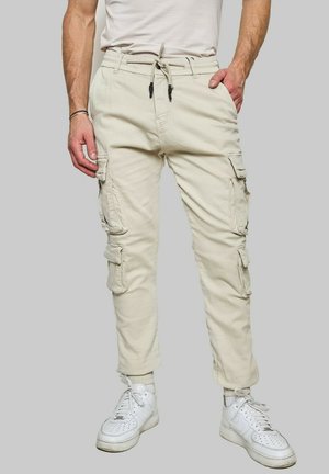 Pantalons cargo beiges avec une taille élastique, fermeture à cordon, plusieurs poches latérales et jambes fuselées. Portés avec des baskets blanches.