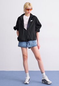 Črn vetrovka z zadrgo spredaj, bela majica, jeans kratke hlače in sivi superge. Model nosi sončna očala s pomarančno barvno lečo.