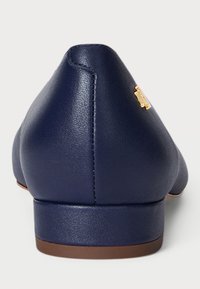 Sapatilha azul-marinho com bico arredondado, feita de couro sintético liso. Apresenta um detalhe em metal dourado de um lado e um salto baixo.