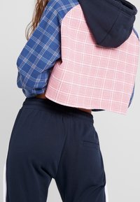 Persona che indossa un hoodie cropped a quadri bicolore con cappuccio e maniche blu navy, abbinato a pantaloni jogger blu navy con tasca laterale.