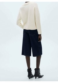 Beige gestrickter Pullover mit umgeschlagenem Kragen, kombiniert mit dunklen Denim-Culottes, getragen mit schwarzen Knöchelschuhen, von hinten gezeigt.