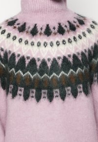 Sweter dziany z wysokim kołnierzem, miękkim różowym korpusem oraz wzorzystą karczkiem z geometrycznymi wzorami w czerni, bieli i brązie.