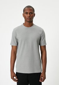 Harmaa lyhythihainen t-paita, jossa on ribbaustekstuuri, pyöreä pääntie ja ompeluyksityiskohtia. Yhdistetty mustiin housuihin, esillä valkoista taustaa vasten.