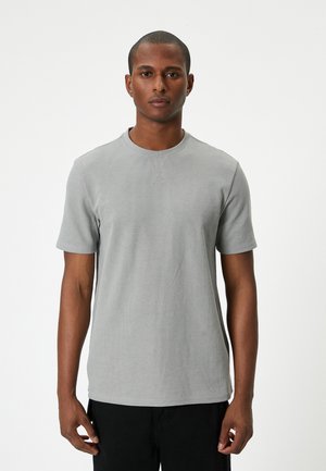Grå t-shirt med korte ærmer og ribbet tekstur, rund halsudskæring og syningsdetaljer. Båret med sorte bukser, præsenteret mod en hvid baggrund.