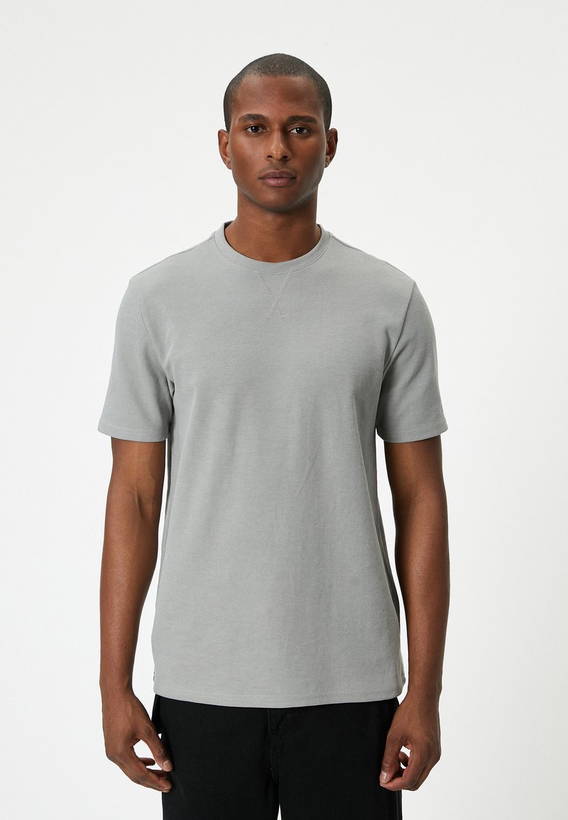 Harmaa lyhythihainen t-paita, jossa on ribbaustekstuuri, pyöreä pääntie ja ompeluyksityiskohtia. Yhdistetty mustiin housuihin, esillä valkoista taustaa vasten.