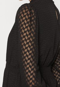 Blouse noire avec des manches en dentelle à motif zigzag transparent et une taille froncée, montrant le haut du dos et la zone des épaules.