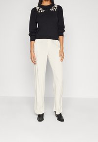 Femme portant un pull à manches longues noir avec broderie florale blanche, un pantalon large crème et des bottines noires, debout devant un fond blanc.