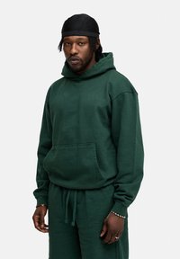 Grüne Kapuzenjacke aus weichem Material, mit einer Vordertasche, weiten Bündchen und einem lässigen Schnitt. Dazu passende Jogginghosen.