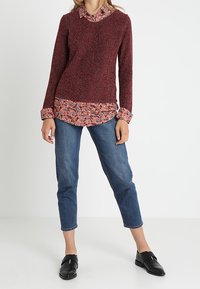 Maglione rosso con texture e colletto floreale indossato sopra una camicia botton-down chiara con motivi floreali, abbinato a jeans blu cropped e scarpe slip-on nere.