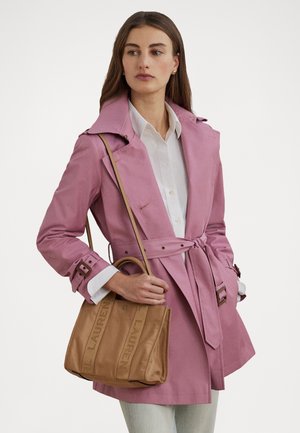 Vrouw draagt een roze trenchcoat met ceintuur en een wit overhemd, houdt een beige Lauren tote bag met schouderriem over haar arm.