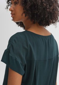 Donker teal blouse met korte, licht geribbelde mouwen, ronde halslijn en een gladde textuur; heeft een naad over de rug.