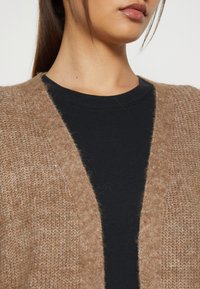 Beżowy sweter na drutach z otwartym przodem, z miękką teksturą i ściągaczami na krawędziach, nałożony na czarną bluzkę z okrągłym dekoltem.