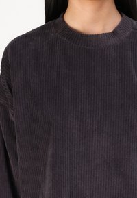Sötét szürke kordbársony felső, bordázott textúrával, kerek nyakkivágással és ejtett vállakkal, közeli felvétel, amely kiemeli a szövet részleteit.
