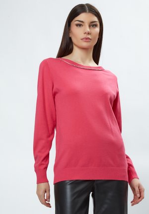 Roze trui met lange mouwen, geribbelde manchetten en zoom, met een bootnek versierd met kleine metalen accenten. Gemaakt van zachte stof.