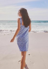 Blaues ärmelloses Kleid mit Blattmuster, knielang, aus leichtem Stoff. Im Hintergrund sind Strand, Meereswellen und sandiges Ufer zu sehen.