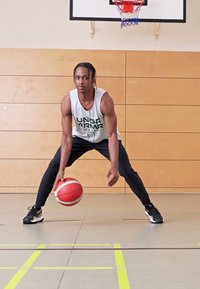 Basketball player in a white tank top and black pants dribbles a red and white basketball on a gym floor with yellow lines and wooden walls.