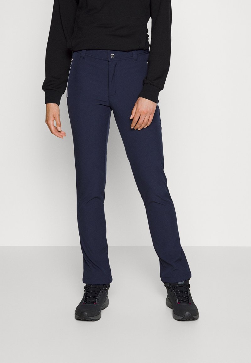 Personne portant un pantalon slim bleu foncé et des chaussures de randonnée noires, debout devant un fond blanc uni.