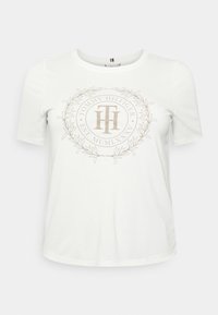 White cotton t-shirt with short sleeves, featuring a beige circular logo with "Tommy Hilfiger" and laurel design in the center.