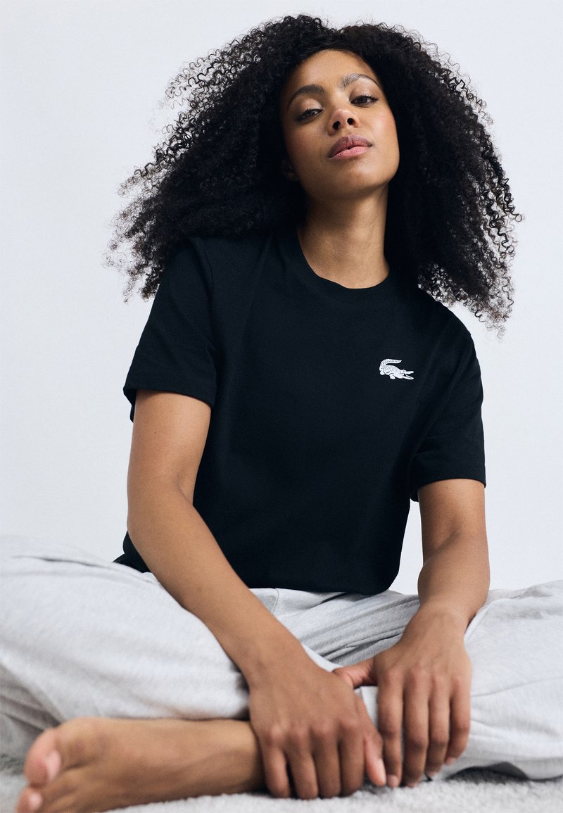 T-shirt de algodão preto com decote redondo e mangas curtas, apresentando um logo de crocodilo bordado em branco no lado esquerdo do peito.