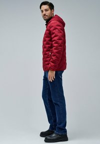 Hombre con chaqueta de plumas roja con capucha, jeans oscuros y botas negras, de pie de lado contra un fondo gris liso.