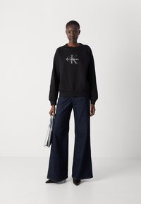 Čierna mikina s logom, skombinovaná so širokými nohavicami z tmavého denimu. Modelka drží striebornú kabelku a má na sebe čierne topánky s špicatou špičkou.
