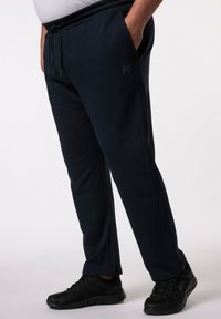 Pantalones de chándal azul marino con cinturilla elástica, bolsillos laterales y un detalle de logotipo. Llevados con zapatillas deportivas negras. Textura de tela suave.